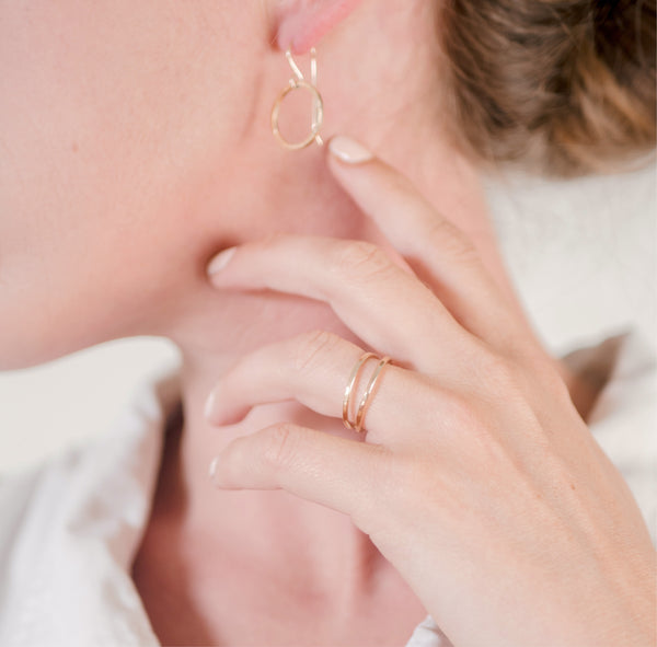 Close up of a woman's hands and earlobe in front of a white wall wearing a white shirt and handcrafted circle dangle earrings and double band vermeil ring by Krista Knickerbocker Designs.