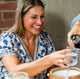 Woman laughing and holding crystal glass as a friend's hand fills her drink.  She is wearing a floral dress and layered gold filled elongated link everyday chain necklaces by Krista Knickerbocker Designs. 