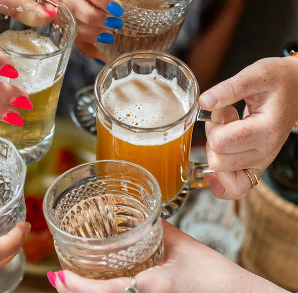 Close up of a group of woman's manicured hands holding glass beer glasses in a "cheers" motion.  Hand in focus is wearing a handmade Krista Knickerbocker Designs gold vermeil double band ring.