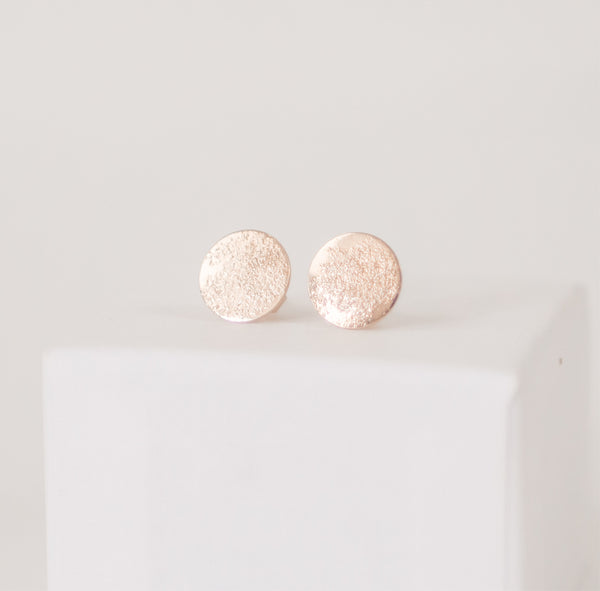 Close up of a pair of shiny, textured circular handcrafted gold post earrings on a white box by Krista Knickerbocker Designs.