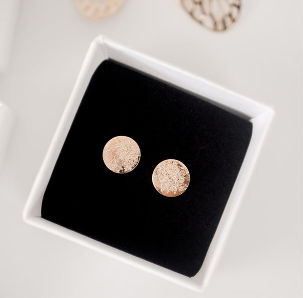 Close up image of shiny, textured gold handcrafted circle stud earrings on a black velvet backing in a white cardboard jewelry box by Krista Knickerbocker Designs.