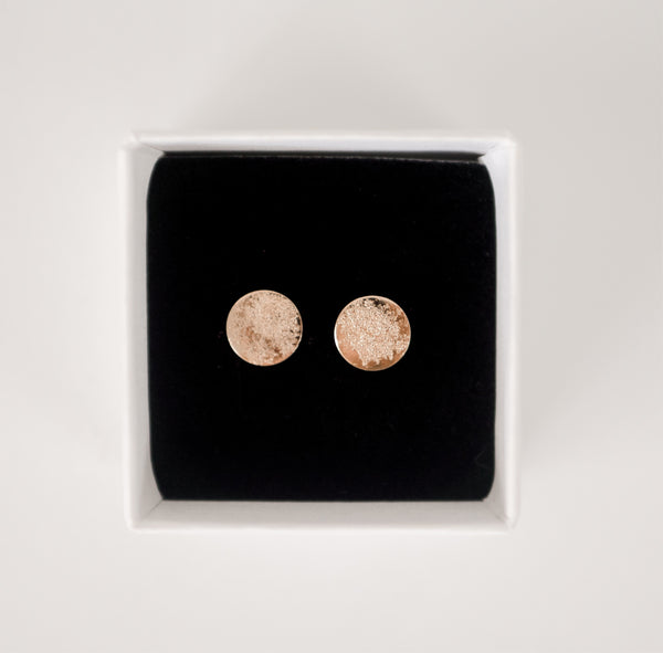 Close up image of shiny, textured gold handcrafted circle stud earrings on a black velvet backing in a white cardboard jewelry box by Krista Knickerbocker Designs.