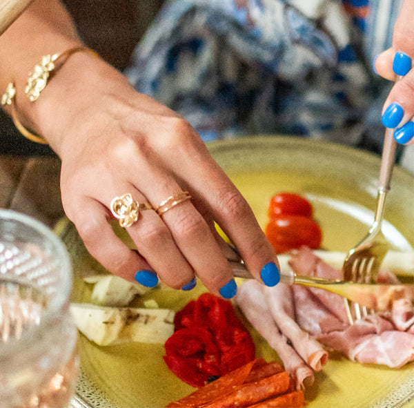 A woman's hands with a blue manicure holding brass silverware and cutting  a plate of Italian lunch meats while wearing handcrafted gold vermeil rings by Krista Knickerbocker Designs.