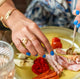 A woman's hands with a blue manicure holding brass silverware and cutting  a plate of Italian lunch meats while wearing handcrafted gold vermeil rings by Krista Knickerbocker Designs. 
