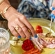 A woman's hands with a blue manicure holding brass silverware and cutting  a plate of Italian lunch meats while wearing handcrafted gold vermeil rings by Krista Knickerbocker Designs.