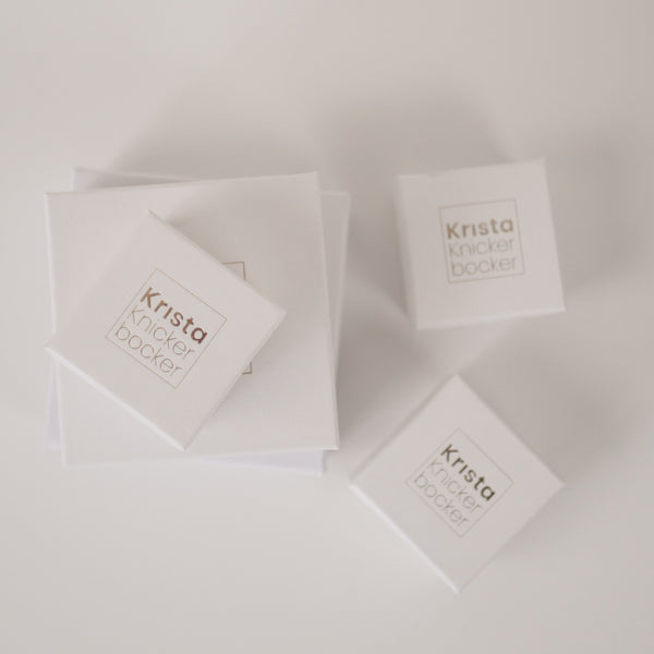 White cardboard jewelry boxes with gold foil embossing that reads Krista Knickerbocker to show packaging that the company's handcrafted jewelry comes in. 