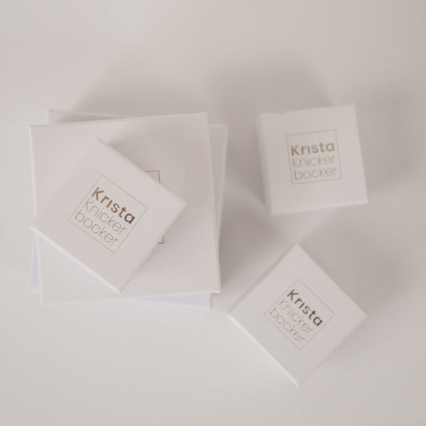 White cardboard jewelry boxes with gold foil embossing that reads Krista Knickerbocker to show packaging that the company's handcrafted jewelry comes in.