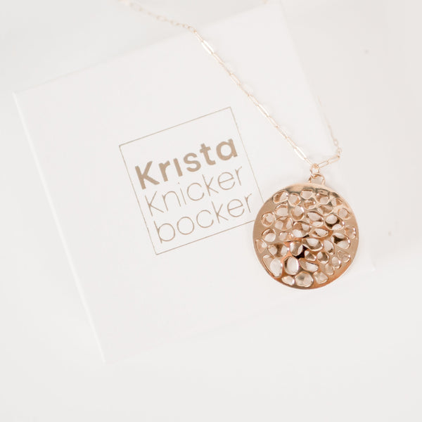 White cardboard jewelry box with gold embossed  name Krista Knickerbocker and a handcrafted, layered, 14 karat gold vermeil calm waters necklace by the designer draped over the box.