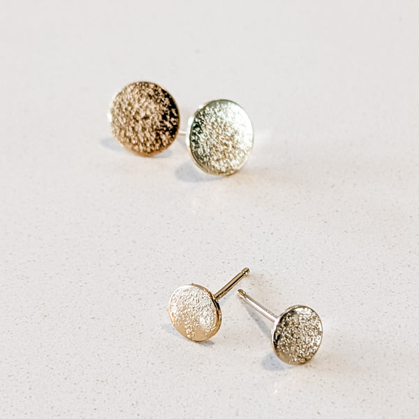 Close up of two sizes, large and petite, of textured gold handcrafted circle "stardust" stud earrings by Krista Knickerbocker Designs.  Earrings are on a textured white surface.
