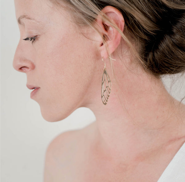 Woman's profile in front of a white wall wearing a white top and gold dragonfly wing dangle earrings by Krista Knickerbocker Designs. 
