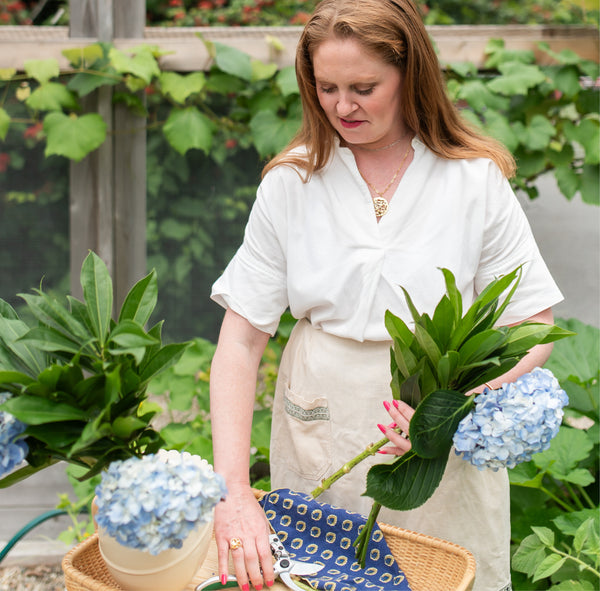 Beautiful woman arranging blue hydrangeas in a garden wearing a white shirt and tan apron and 14k gold water reflection handcrafted necklace by Krista Knickerbocker Designs. 