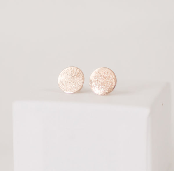 Close up of a pair of shiny, textured circular handcrafted gold post earrings on a white box by Krista Knickerbocker Designs. 