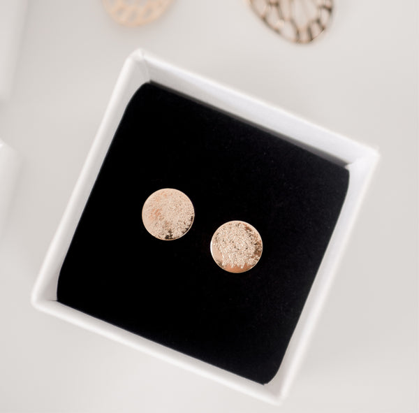 Close up image of shiny, textured gold handcrafted circle stud earrings on a black velvet backing in a white cardboard jewelry box by Krista Knickerbocker Designs. 