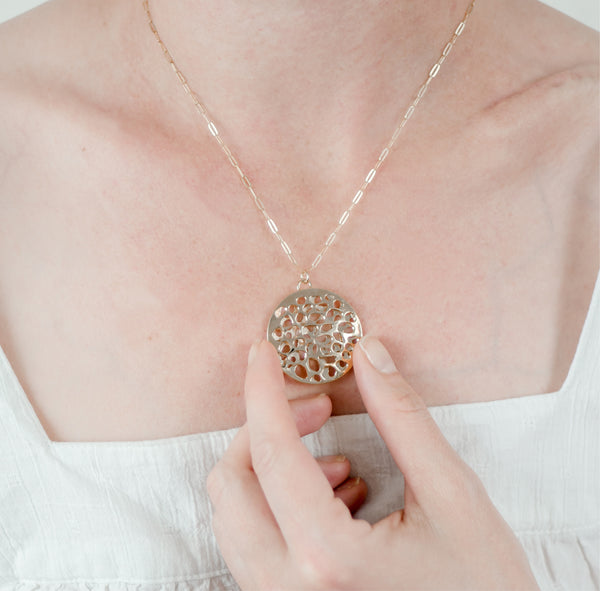 Zoom in of a woman wearing a white shirt and gently holding a large circle handcrafted gold pendant inspired by light reflecting off of water and crafted by Krista Knickerbocker Designs. 