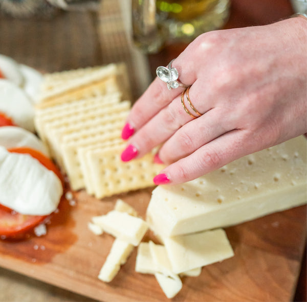 Woman's hand with pin manicure reaching towards a cheese plate wearing Krista Knickerbocker Designs' handcrafted gold vermeil double band ring and sterling silver cluster ring. 