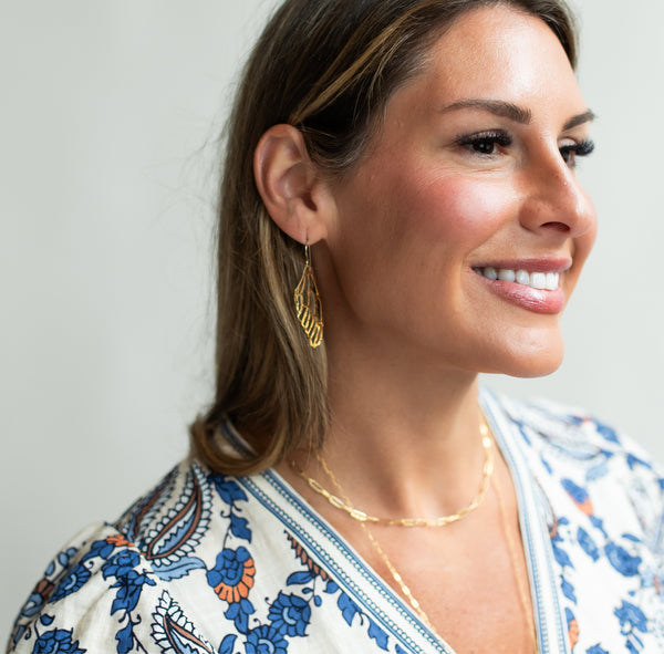 Beautiful smiling woman in a floral dress in front of a white wall wearing gold chain necklaces and gold dangle dragonfly wing earrings by Krista Knickerbocker Designs. 
