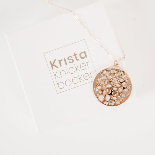 White cardboard jewelry box with gold embossed  name Krista Knickerbocker and a handcrafted, layered, 14 karat gold vermeil calm waters necklace by the designer draped over the box. 