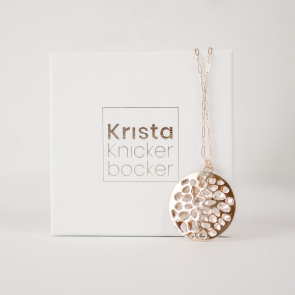 White cardboard jewelry box with gold embossed  name Krista Knickerbocker and a handcrafted, layered, 14 karat gold vermeil calm waters necklace by the designer draped over the box. 