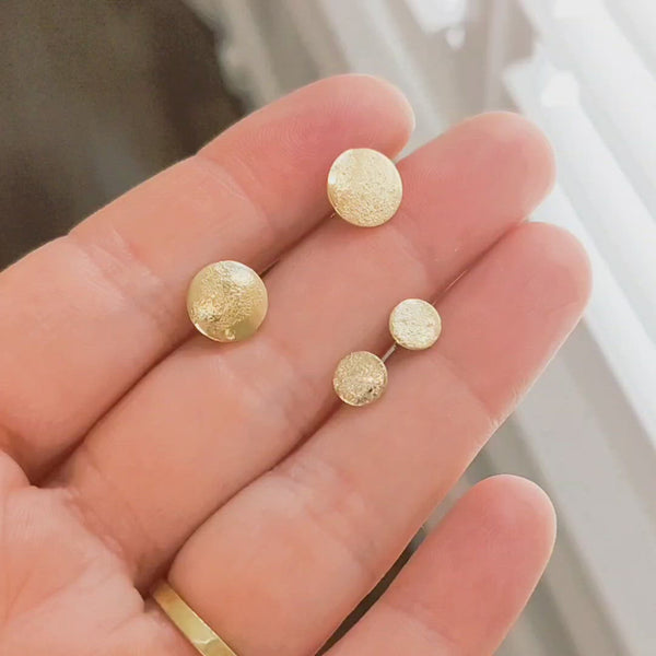 Hand holding two sizes, large and petite, of shiny gold handmade textured "stardust" stud earrings by Krista Knickerbocker Designs.  The hand is moving slowly showing how the light reflects off of the pieces. 